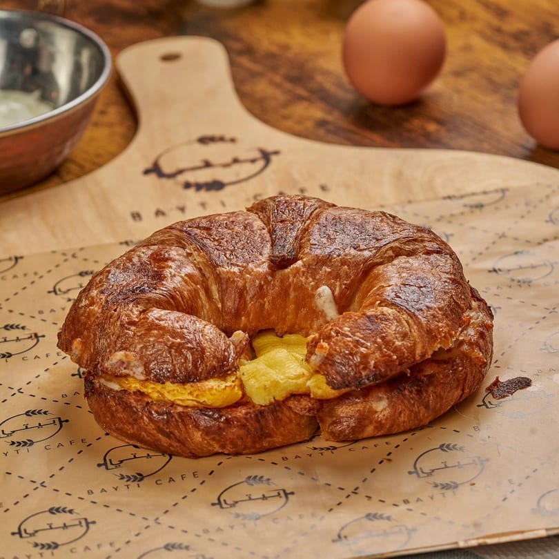 Golden croissant sandwich with scrambled eggs on Bayti Cafe branded paper, with eggs and bowl in background.