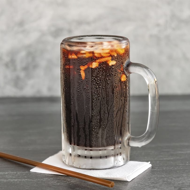 A cold, dark beverage in a condensation-covered glass mug with ice and a straw.