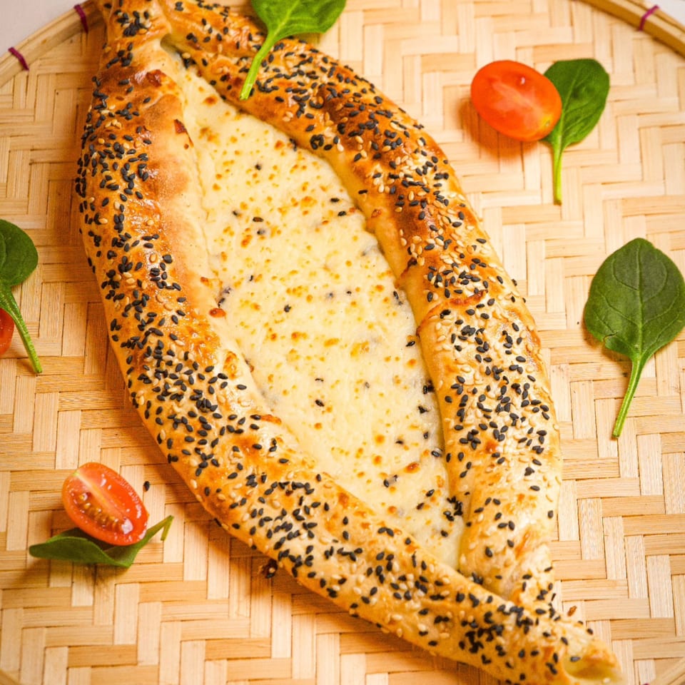 Lebanese cheese manakish, boat-shaped, topped with sesame seeds, served on a woven mat with spinach and tomatoes.