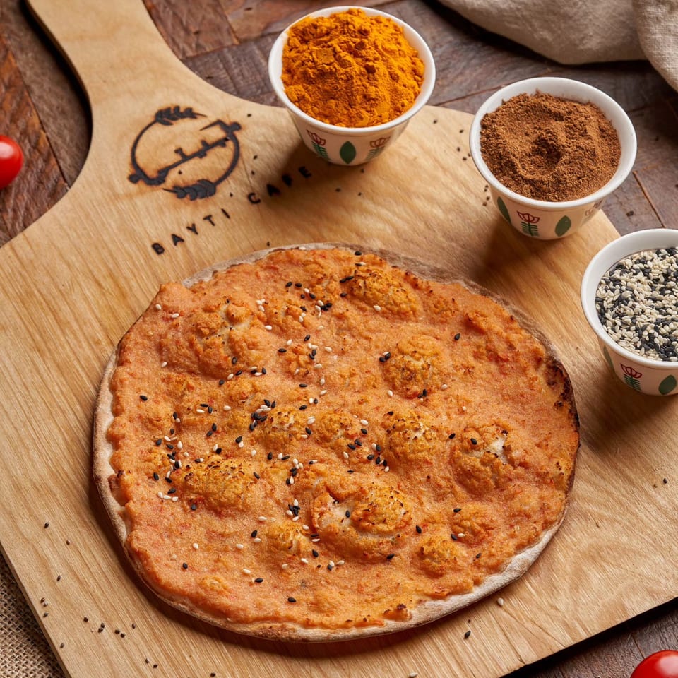 Lebanese flatbread with orange topping and sesame seeds, alongside bowls of spices on a wooden board.