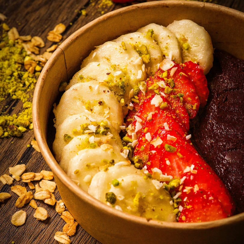 A Bayti Cafe acai bowl with banana, strawberries, and pistachios on a wooden table.