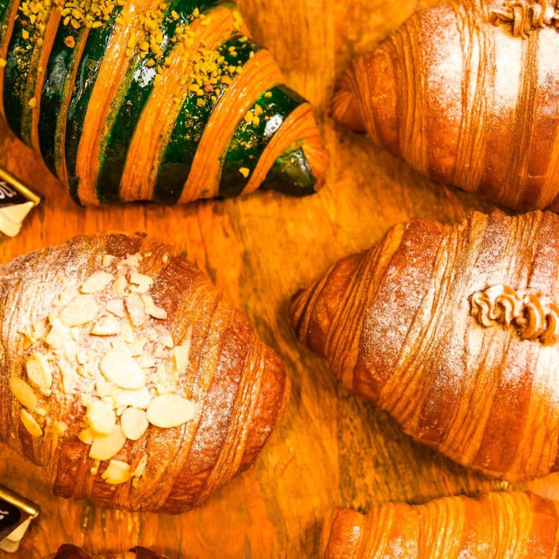 Assortment of croissants, including one with green stripes and pistachios, on a wooden surface.
